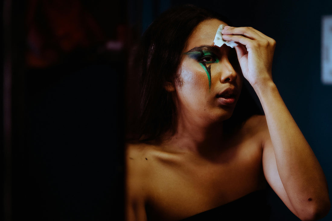 Person removing bold green eye makeup with tissue in dimly lit room