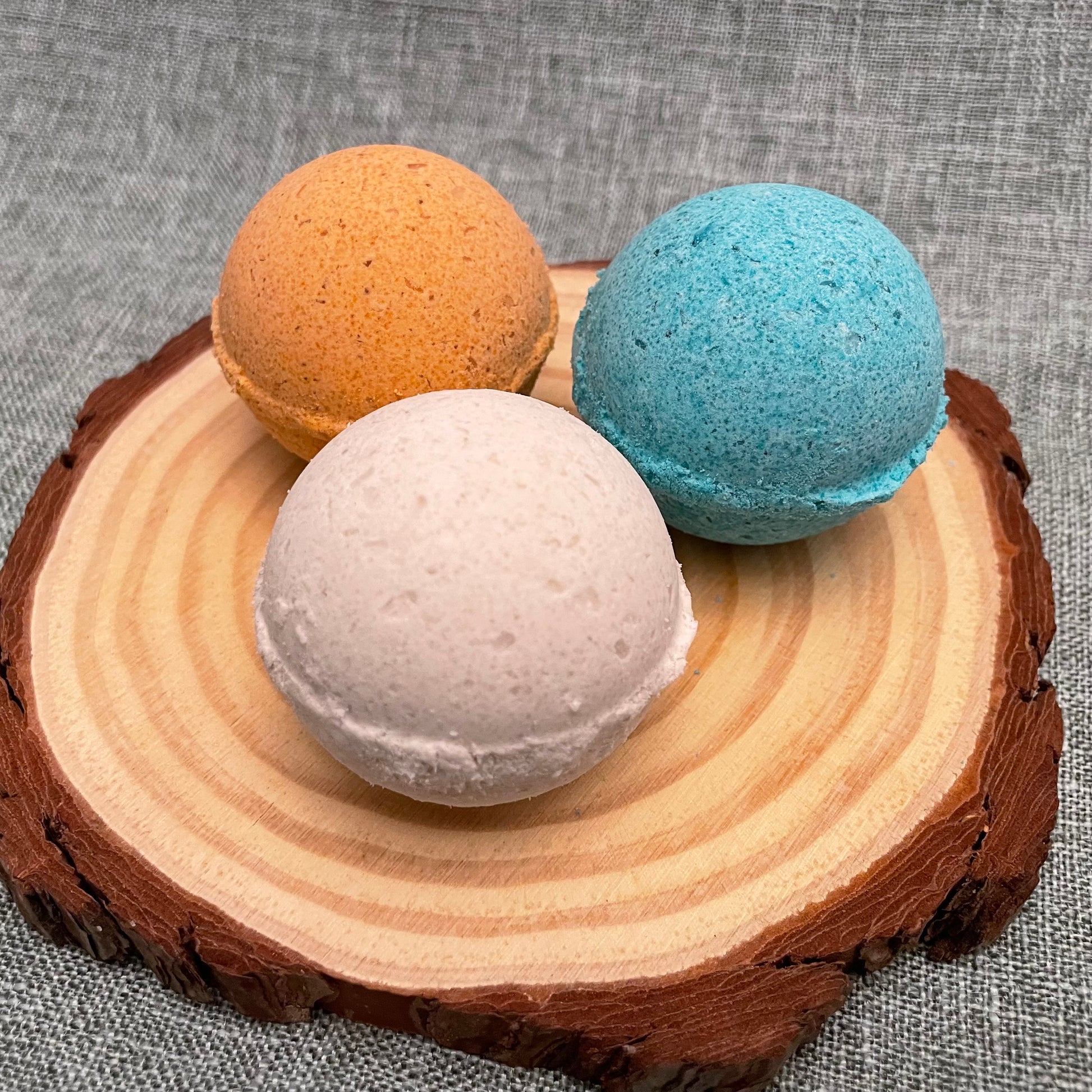 Three bath bombs in brown, white, and blue on a wooden block with a gray fabric background.
